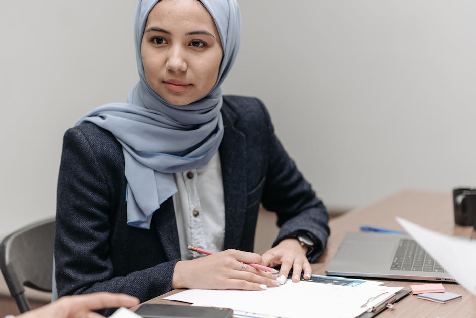 A woman in a hijab working at an office desk with a laptop and documents.